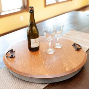 Wine bottle and two glasses on a wooden round tray with metal handles, placed on a dark wooden table with a woven beige table runner.