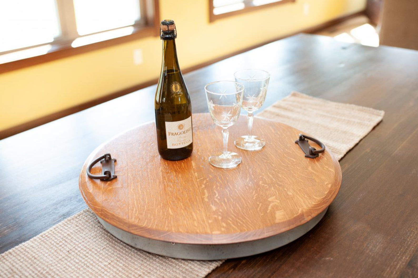 Wine bottle and two glasses on a wooden round tray with metal handles, placed on a dark wooden table with a woven beige table runner.