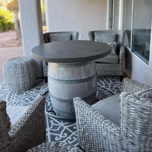 Outdoor patio setup with wicker chairs, a barrel-base table with a dark round top, woven ottoman, and patterned rug on tiled floor.