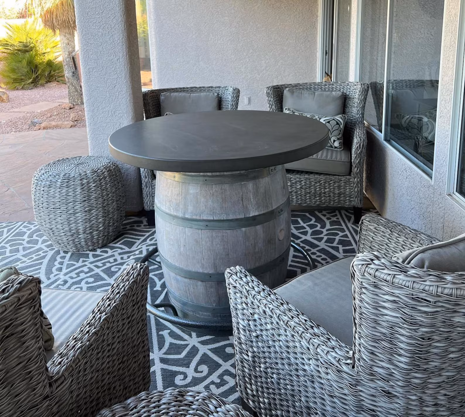 Outdoor patio setup with wicker chairs, a barrel-base table with a dark round top, woven ottoman, and patterned rug on tiled floor.