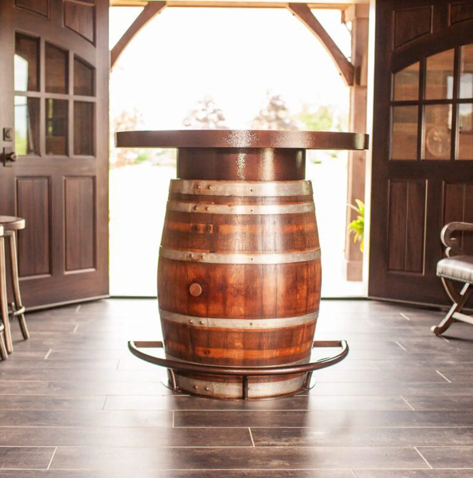 Wooden barrel repurposed as a standing table with metal footrest, placed indoors near open doors with sunlight outside.