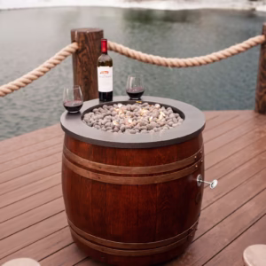 Wine barrel fire pit on a wooden deck by a lake, topped with lava rocks, a wine bottle, and two glasses, surrounded by snow.