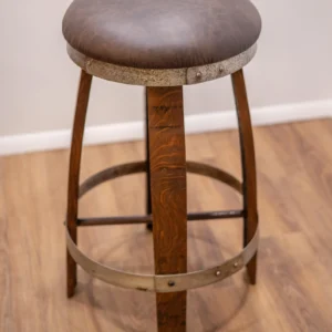 Rustic bar stool with a round brown leather cushion, wooden legs, and metal bands, placed on a wood floor against a plain wall.