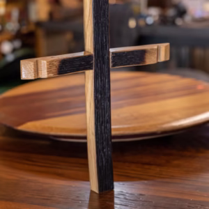 A wooden cross with charred black and natural wood finish stands upright on a polished wooden table with a blurred background.